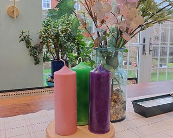 Op de afbeelding: Drie kaarsen op een houten voet. De kaarsen zijn roze, groen en paars. De kaarsen staan op een witte tafelkleed met een geruit patroon.