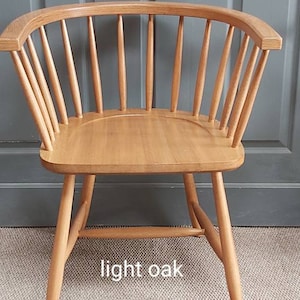 Op de afbeelding: Een stoel van licht eikenhout met een gebogen rugleuning en een latten ontwerp. De stoel staat op een lichtbruin tapijt en heeft een houten bord met de tekst "CHAIR SUPPLY" ervoor.