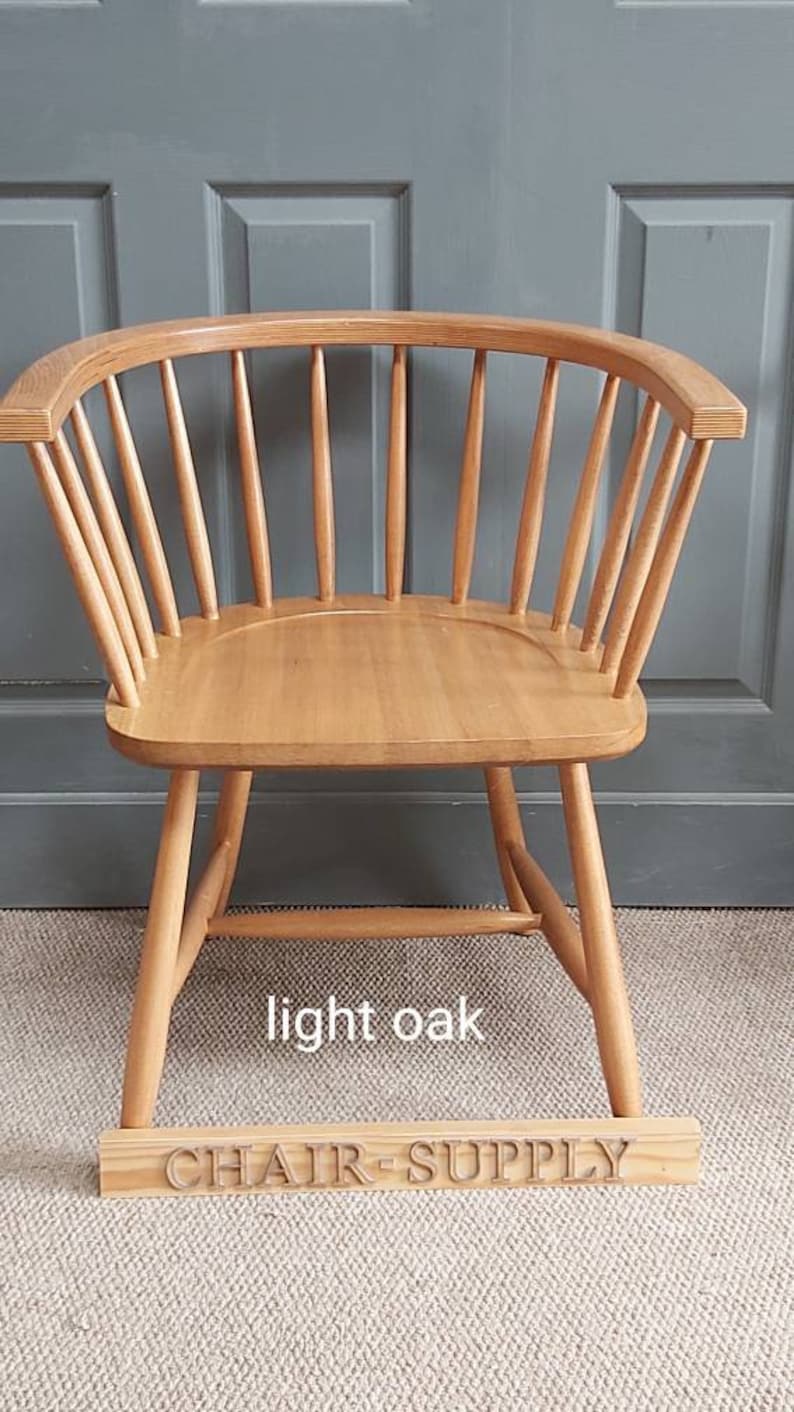 Op de afbeelding: Een stoel van licht eikenhout met een gebogen rugleuning en een latten ontwerp. De stoel staat op een lichtbruin tapijt en heeft een houten bord met de tekst "CHAIR SUPPLY" ervoor.