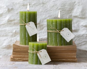 Op de afbeelding: Drie olijfgroene, geribbelde stompkaarsen met brandende lonten. Elke kaars is omwikkeld met een dun beige touwtje en voorzien van een klein wit rechthoekig papieren label met de tekst PILLAR CANDLE en een cirkelvormig logo-ontwerp.