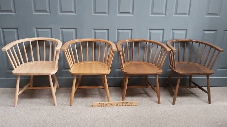 Op de afbeelding: Vier houten stoelen met een gebogen rugleuning en een spindelontwerp. De stoelen zijn in verschillende stadia van vlekken, van licht tot donker. De stoelen zijn in een rij op een houten vloer voor een grijze muur geplaatst. De tekst "CHAIR-SUPPLY" is zichtbaar op een houten bord voor de stoelen.