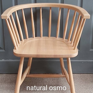 Op de afbeelding: Een stoel van natuurlijk hout met een gebogen rugleuning en een brede zitting. De stoel is gemaakt van massief hout en heeft een natuurlijke afwerking. De stoel staat op een tapijt met een houten bord waarop "CHAIR-SUPPLY" en "natural osmo" staat.