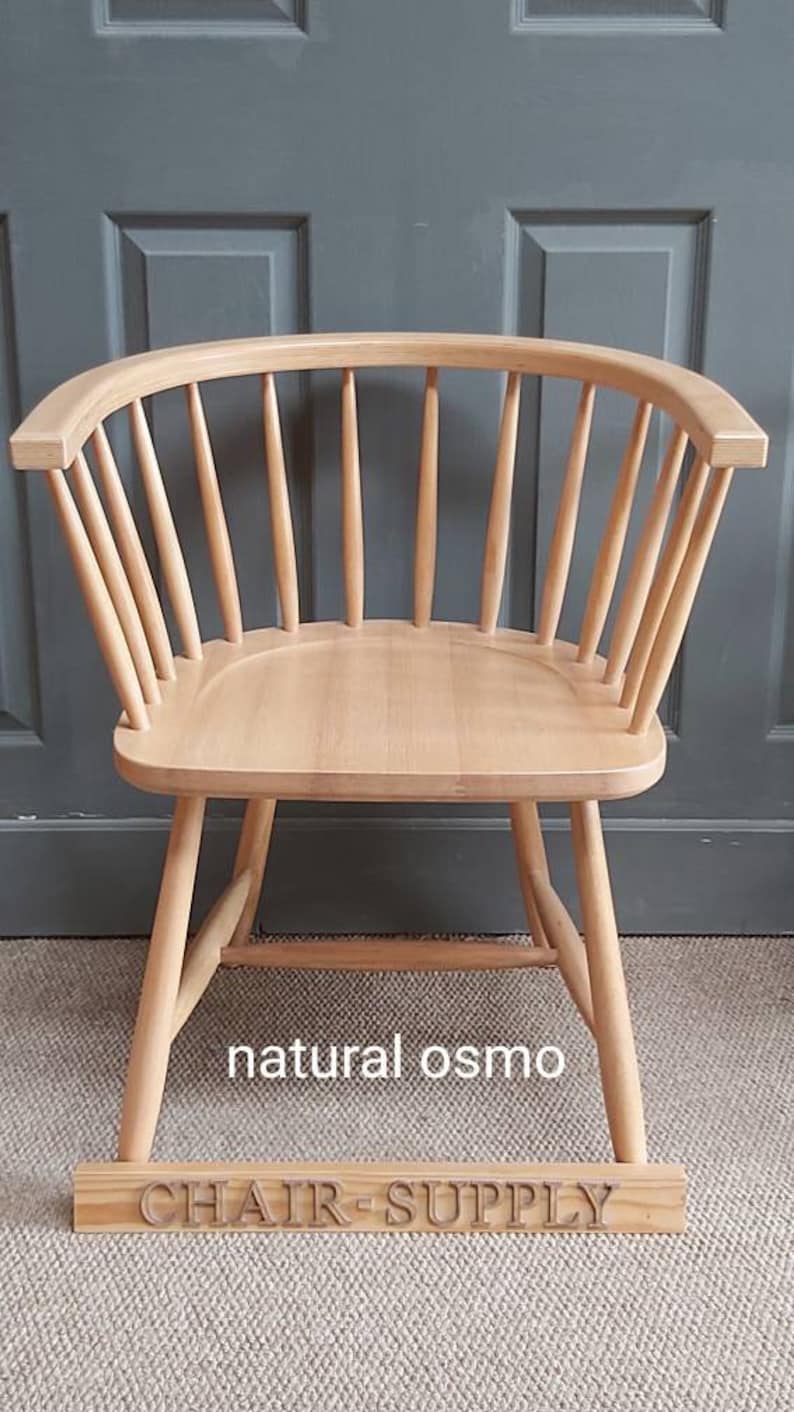 Op de afbeelding: Een stoel van natuurlijk hout met een gebogen rugleuning en een brede zitting. De stoel is gemaakt van massief hout en heeft een natuurlijke afwerking. De stoel staat op een tapijt met een houten bord waarop "CHAIR-SUPPLY" en "natural osmo" staat.