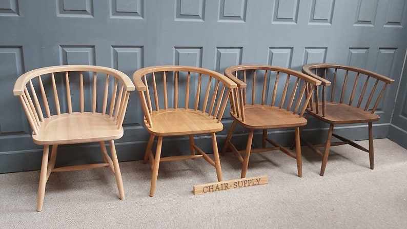 Op de afbeelding: Vier houten stoelen met een gebogen rugleuning en een spindelrug. De stoelen zijn in verschillende houttinten, van licht tot donker. De stoelen zijn in een rij op een tapijt gelegd. Een bord met de tekst "CHAIR-SUPPLY" staat op de voorgrond.
