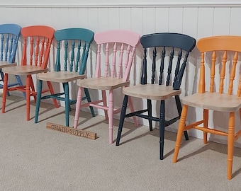 Op de afbeelding: Zes kleurrijke houten stoelen met spijlenrug en natuurhouten zittingen. De stoelen zijn geschilderd in blauw, koraal, blauwgroen, roze, marineblauw en oranje. De tekst "CHAIR SUPPLY" staat op een houten bord.