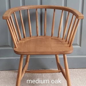 Op de afbeelding: Een houten stoel met een gebogen rugleuning en een spindelrug. De stoel is gemaakt van middelgrote eik en heeft een natuurlijke afwerking. De stoel staat op een tapijt voor een grijze deur. De stoel is gelabeld met "middelgrote eik" en "CHAIR SUPPLY".