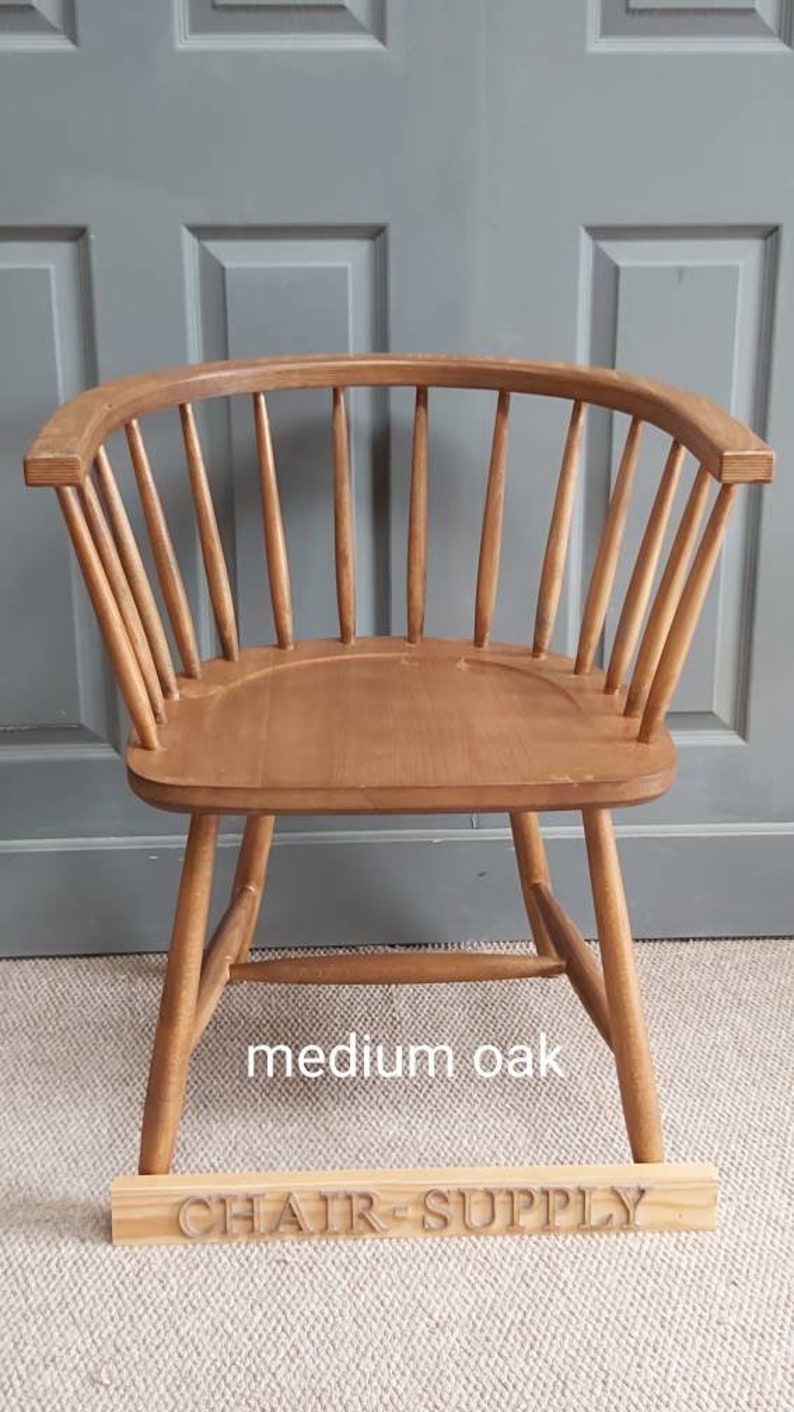 Op de afbeelding: Een houten stoel met een gebogen rugleuning en een spindelrug. De stoel is gemaakt van middelgrote eik en heeft een natuurlijke afwerking. De stoel staat op een tapijt voor een grijze deur. De stoel is gelabeld met "middelgrote eik" en "CHAIR SUPPLY".