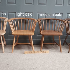 Op de afbeelding: Vier houten stoelen in verschillende eikenhouten tinten, van links naar rechts: natuurlijke osmo, lichte eik, middelste eik en donkere eik. De stoelen zijn allemaal in dezelfde stijl, met een gebogen rugleuning en een brede zitting. De stoelen zijn in een rij op een lichtbruine vloer geplaatst, met een grijze muur erachter. De tekst "CHAIR-SUPPLY" is gedrukt op een houten bord voor de stoelen.