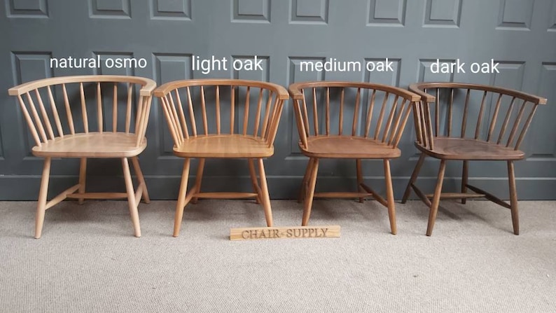 Op de afbeelding: Vier houten stoelen in verschillende eikenhouten tinten, van links naar rechts: natuurlijke osmo, lichte eik, middelste eik en donkere eik. De stoelen zijn allemaal in dezelfde stijl, met een gebogen rugleuning en een brede zitting. De stoelen zijn in een rij op een lichtbruine vloer geplaatst, met een grijze muur erachter. De tekst "CHAIR-SUPPLY" is gedrukt op een houten bord voor de stoelen.