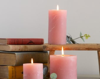 Op de afbeelding: Drie roze kaarsen met een getextureerd oppervlak. De kaarsen zijn aangestoken en staan op een houten plank met een stapel boeken eronder.