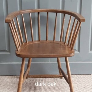 Op de afbeelding: Een stoel van donker eikenhout met een gebogen rugleuning en een eenvoudig ontwerp. De stoel staat op een lichtbruin tapijt voor een grijze deur. De tekst "CHAIR SUPPLY" is zichtbaar op een houten bord voor de stoel.