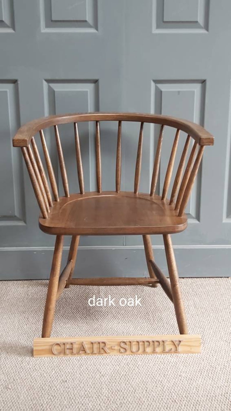 Op de afbeelding: Een stoel van donker eikenhout met een gebogen rugleuning en een eenvoudig ontwerp. De stoel staat op een lichtbruin tapijt voor een grijze deur. De tekst "CHAIR SUPPLY" is zichtbaar op een houten bord voor de stoel.