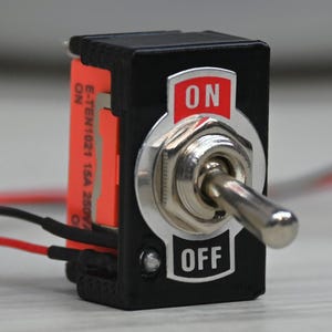 Op de afbeelding: Een zwarte tuimelschakelaar met een zilveren hendel en rode en zwarte draden. De schakelaar heeft de labels "ON" en "OFF". Aan de zijkant is een oranje label zichtbaar. De schakelaar staat op een lichtgekleurd oppervlak.
