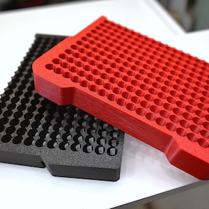 May include: Two plastic trays, one red and one black, are displayed on a white surface. Both trays have a grid of circular holes. The red tray is on top, partially obscuring the black tray. The trays appear to be designed for organizing small items.