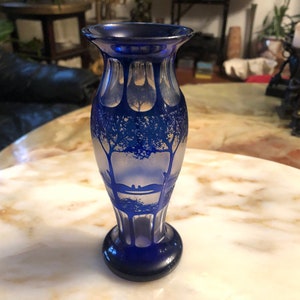 May include: A cobalt blue glass vase with a frosted design of trees and a bridge. The vase has a rounded body and a flared lip.