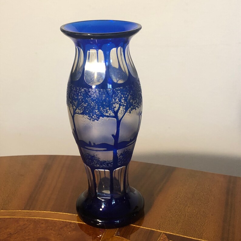 May include: A cobalt blue glass vase with a frosted design of trees and a landscape. The vase has a rounded body and a flared lip.