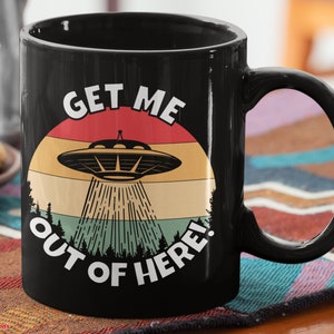 May include: Black coffee mug with a retro-style graphic of a UFO and the text "Get Me Out Of Here!"