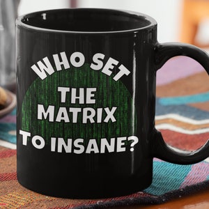 May include: Black ceramic mug with the text "WHO SET THE MATRIX TO INSANE?" in white. The text is over a green background with a matrix-style digital rain pattern. The mug has a curved handle.