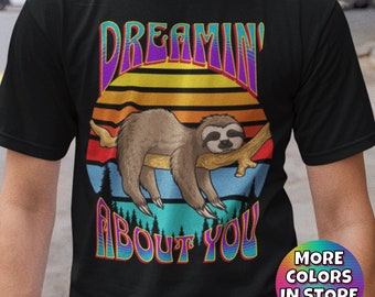 Pode incluir: Uma camiseta preta com um gráfico de estilo retrô de um bicho-preguiça dormindo em um galho. O gráfico é cercado por um pôr do sol com cores de arco-íris e o texto "Dreamin' About You" em uma fonte colorida.
