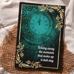 May include: A teal green and gold journal cover with a clock face and intricate details. The text on the cover reads "Ticking away the moments that make up a dull day."