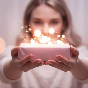 Puede incluir: Una caja de regalo rosa claro sostenida con ambas manos, con luces brillantes que emanan de la parte superior. El fondo presenta luces bokeh suaves y borrosas, creando una atmósfera de ensueño y festiva.
