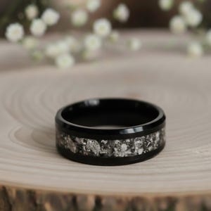 Può includere: Un anello nero con intarsio argentato. L'anello è posto su una superficie di legno, con fiori bianchi sullo sfondo. La fascia dell'anello è nera e l'intarsio è un mix di argento e grigio.