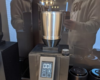 Könnte beinhalten: Eine schwarze Kaffeemühle mit einem digitalen Display, das "00.5" anzeigt. Die Mühle hat einen silbernen Portafilterhalter aus Metall und eine schwarze Basis mit dem aufgedruckten Wort "SPECIALITA".