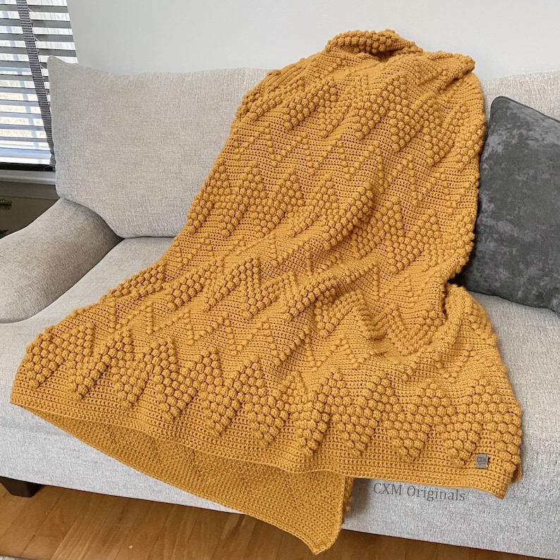 Puede incluir: Una manta de punto de crochet de color amarillo mostaza con un patr&oacute;n texturizado de punto bobble. La manta est&aacute; drapeada sobre un sof&aacute; y presenta el texto "CXM Originals".