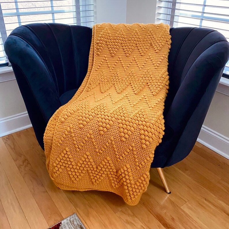 Puede incluir: Una manta de crochet de color amarillo mostaza con un patr&oacute;n texturizado de punto bobble drapeado sobre un sill&oacute;n de terciopelo negro.