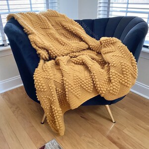 Puede incluir: Una manta de crochet de color amarillo mostaza con un patr&oacute;n de punto de bobble texturizado, drapeada sobre un sill&oacute;n negro con patas doradas.