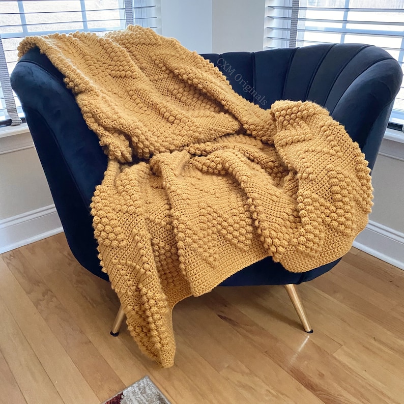 Puede incluir: Una manta de crochet de color amarillo mostaza con un patr&oacute;n de punto de bobble texturizado, drapeada sobre un sill&oacute;n negro con patas doradas.