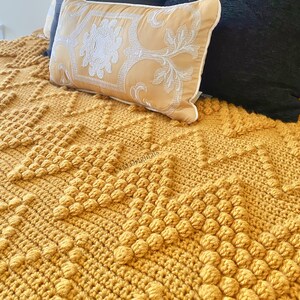 Puede incluir: Una manta de crochet de color amarillo mostaza con un dise&ntilde;o texturizado y abultado. La manta est&aacute; doblada y tiene una almohada decorativa encima.