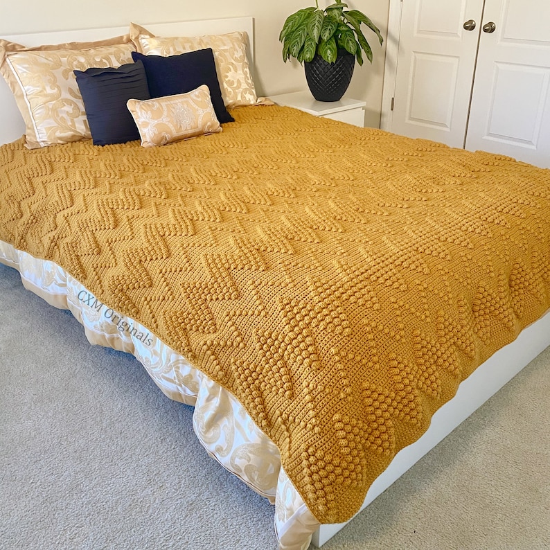 Puede incluir: Una manta de crochet de color amarillo mostaza con un patr&oacute;n texturizado. La manta est&aacute; sobre una cama con fundas de almohada de color blanco y dorado y un marco de cama blanco.