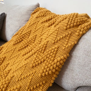 Puede incluir: Una manta de punto de crochet de color amarillo mostaza con un patr&oacute;n de puntadas de pomp&oacute;n texturizado. La manta est&aacute; drapeada sobre un sof&aacute; gris.