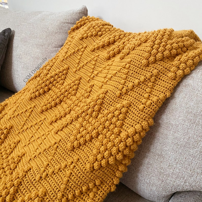 Puede incluir: Una manta de punto de crochet de color amarillo mostaza con un patr&oacute;n de puntadas de pomp&oacute;n texturizado. La manta est&aacute; drapeada sobre un sof&aacute; gris.