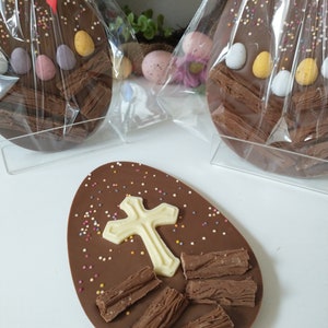 May include: Chocolate Easter eggs, some wrapped in cellophane, are decorated with sprinkles and candy eggs. One egg has a white cross and chocolate pieces. A festive treat for Easter.