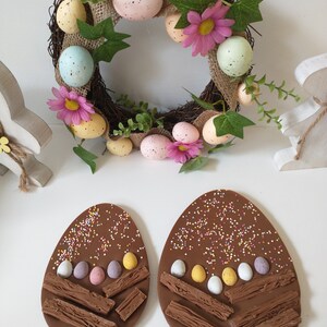 May include: Two chocolate Easter eggs decorated with colourful sprinkles, mini chocolate eggs, and chocolate bar pieces. The eggs are on a white surface.