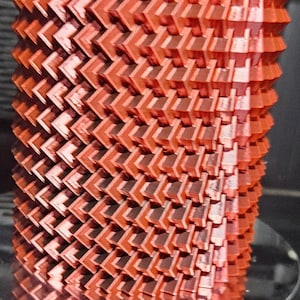 May include: A 3D-printed, coral-colored object with a repeating chevron pattern. The geometric design has a cylindrical shape, with layers of angled, textured surfaces. The object is displayed on a reflective, circular surface.