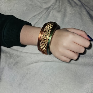 Könnte beinhalten: Ein Set aus drei goldenen und grünen Armreifen mit geometrischem Muster. Die Armreifen sind auf dem Handgelenk einer Person gestapelt.