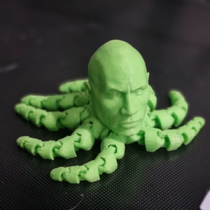 May include: A green 3D printed octopus with a human head. The octopus has eight tentacles and the head has a realistic human face.