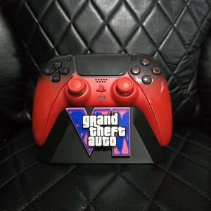 May include: A red and black wireless video game controller featuring a black touchpad, red analogue sticks, and a black faceplate with the Grand Theft Auto VI logo in white text over a blue and pink palm tree graphic, resting on a black stand.