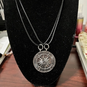 May include: Two matching necklaces with a compass rose pendant. The pendants are silver-colored, circular, and feature a detailed compass design with a central starburst. The necklaces have black cords and are displayed on a black velvet bust.