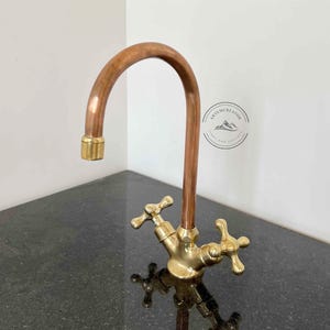 May include: A copper and brass faucet with a curved spout. The faucet has cross-shaped handles and a gold-colored finish on the base and spout end. The faucet is mounted on a dark countertop.