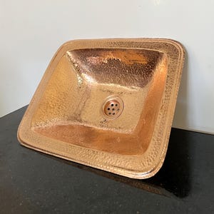 Pode incluir: Uma pia de cobre martelado com uma bacia quadrada e uma borda arredondada. A pia tem um ralo no centro e uma superfície texturizada. O cobre tem uma cor marrom-avermelhada quente e reflete a luz.