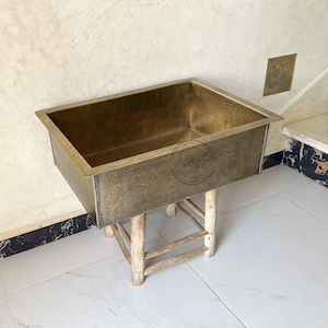 May include: A rectangular, hammered brass sink sits on a light-coloured wooden stand. The sink has a textured surface and is placed against a light beige wall. A small brass plate is mounted on the wall. The floor is white tile.