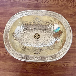 May include: A silver oval-shaped bathroom sink with an ornate design. The sink has a drain in the center and is set against a wood background.