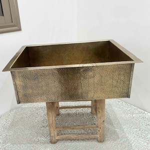 Peut inclure: Un évier rectangulaire en cuivre martelé de couleur dorée, posé sur un support en bois rustique. La surface texturée et les bords angulaires de l'évier sont visibles. Le support a une finition en bois naturel. C'est un élément de cuisine ou de salle de bain.