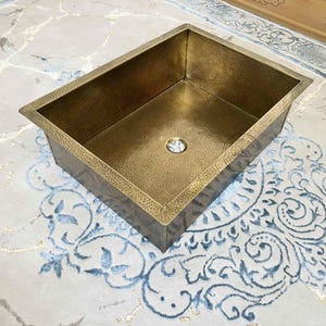 Op de afbeelding: Een rechthoekige, gehamerde koperen spoelbak met een centrale afvoer. De spoelbak heeft een warme, goud-bronzen tint en een getextureerd oppervlak. De spoelbak staat op een patroonkleed met blauwe en witte bloemmotieven. Het is een keuken- of badkamerarmatuur.