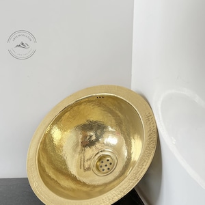 May include: A hammered brass sink with a round shape and a drain in the center. The sink is sitting on a black countertop.