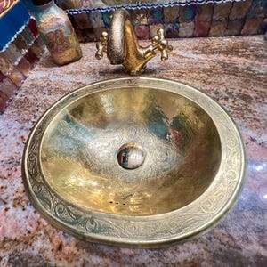 Peut inclure: Un évier ovale en laiton martelé avec un motif floral orné sur le bord et dans le bassin. Un robinet en laiton assorti est monté au-dessus. L'évier est un élément décoratif de salle de bain.